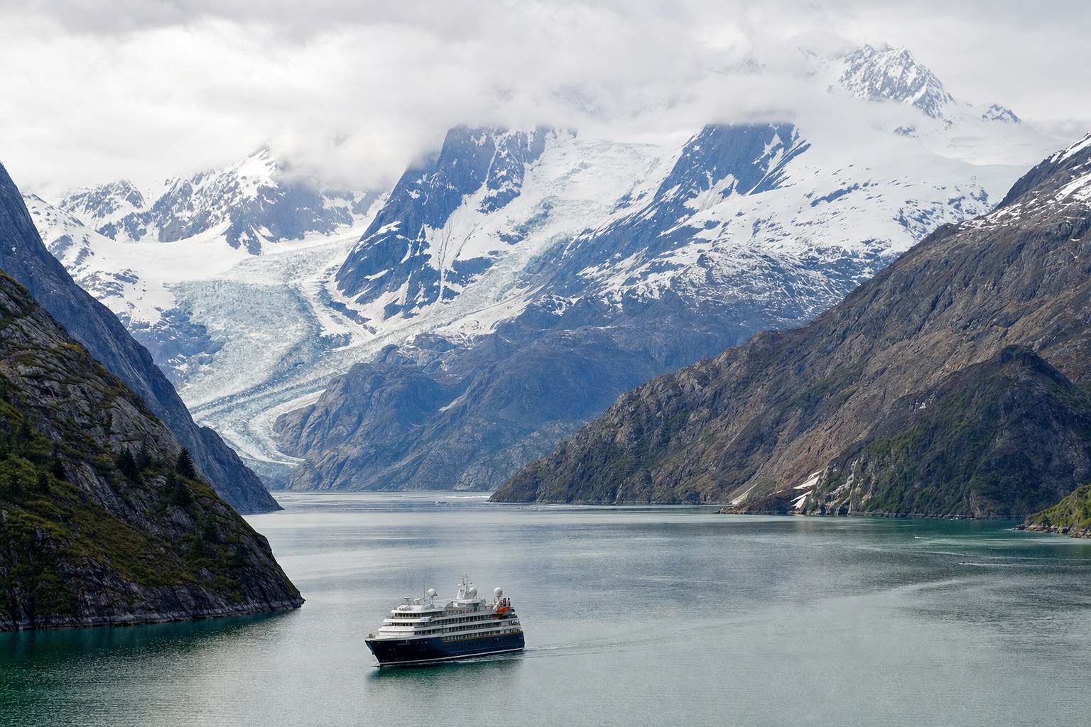 Small cruise ship sailing through an Alaska fjord beneath glacier-covered mountains