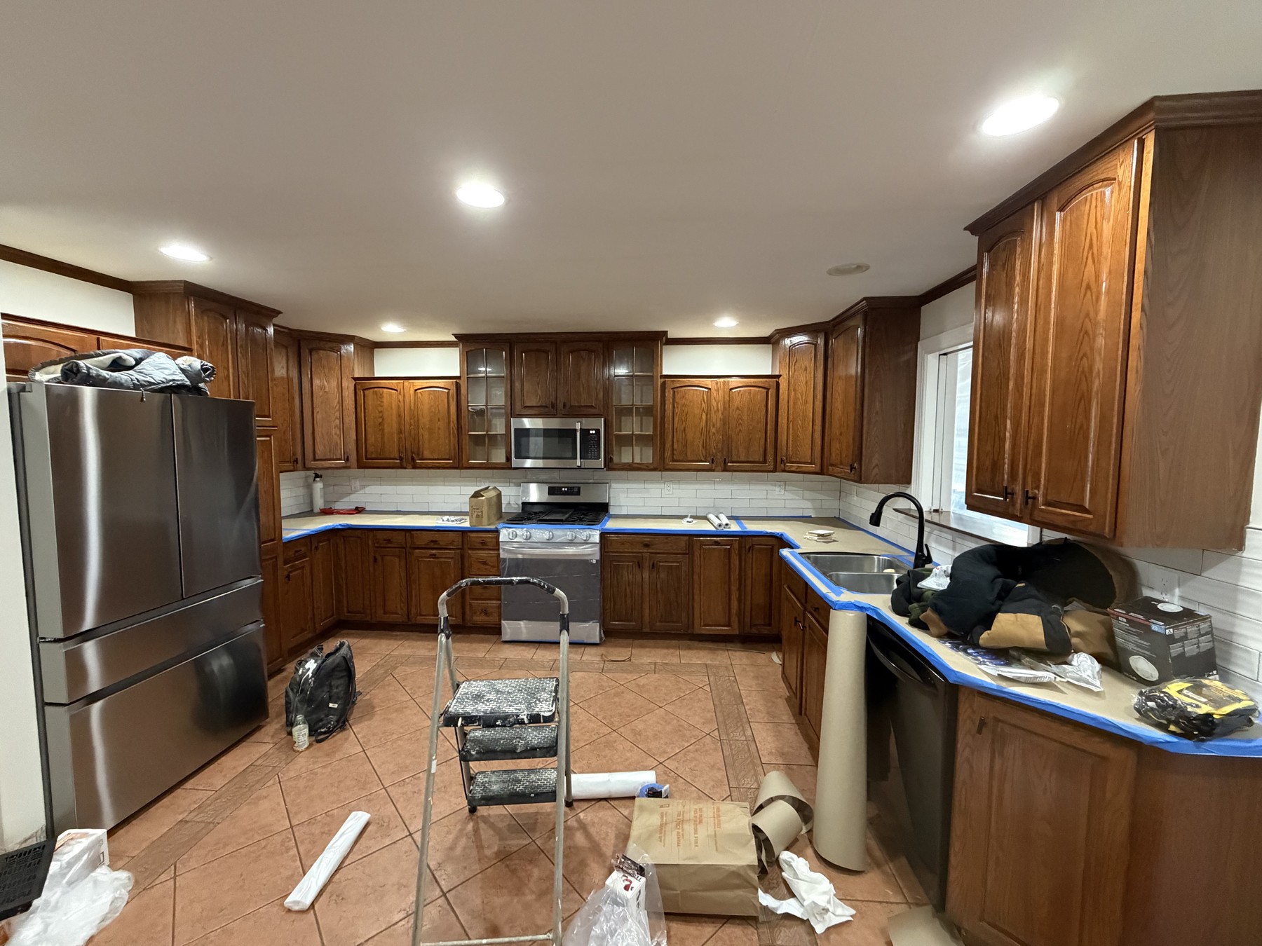 Original wood-finish kitchen cabinets, old tile flooring, taped countertops, and a stepladder during preparation