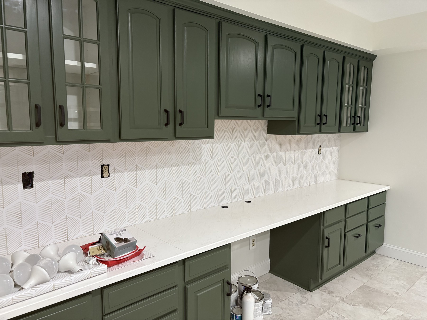 Green repainted cabinets with glass-front doors and black handles above a long counter, with paint and lighting supplies nearby