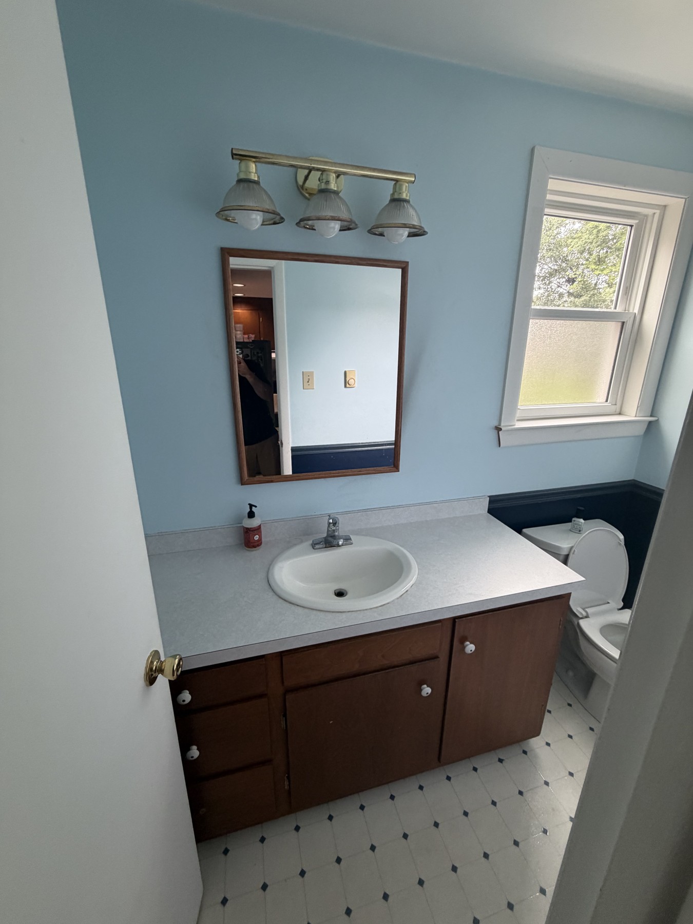Blue powder room before the refresh with a wood vanity, gray countertop, rectangular wood-framed mirror, and older light fixture