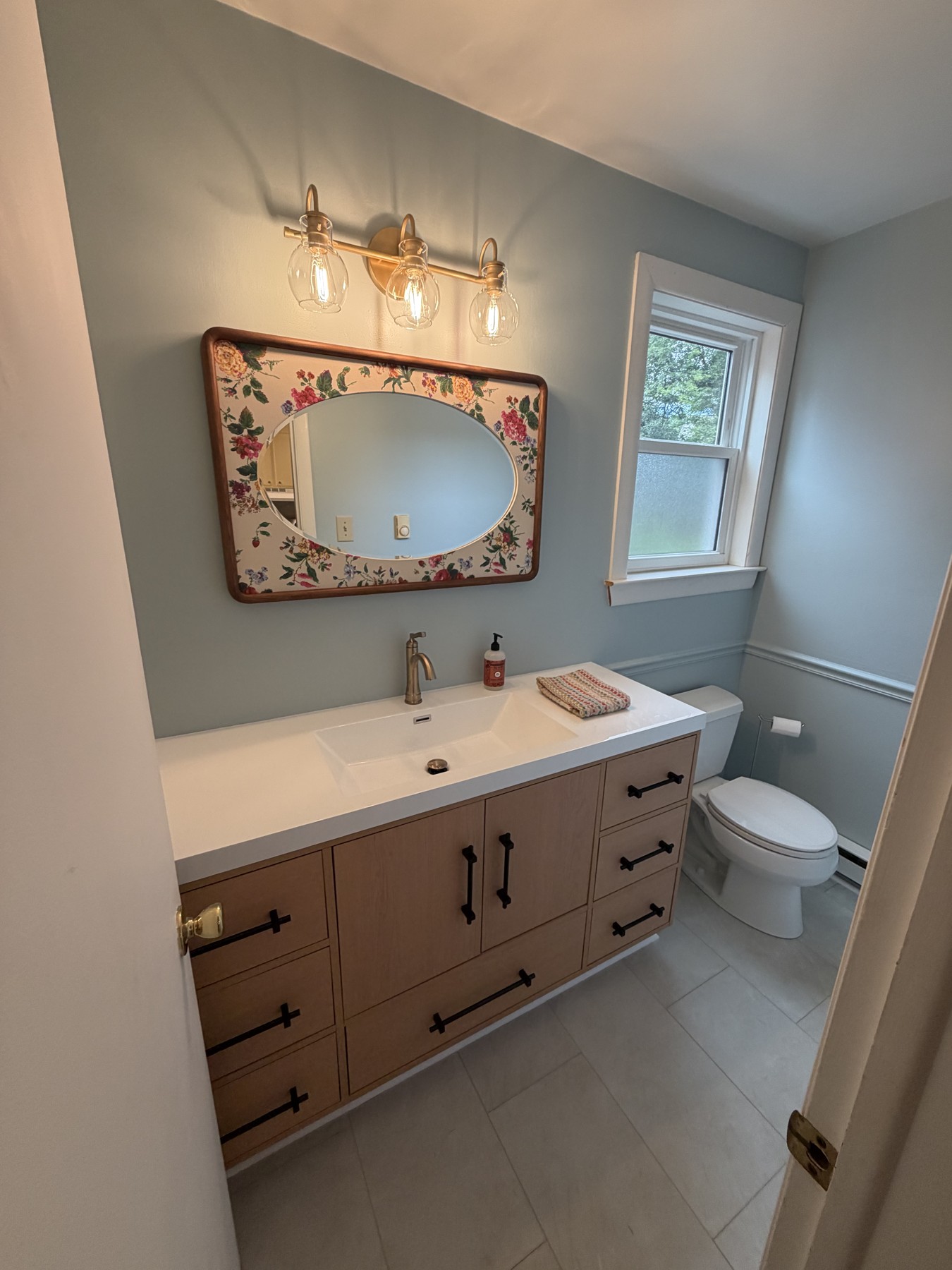 Blue powder room with a light wood vanity, white sink, brass-toned faucet, floral oval mirror, and three-light fixture