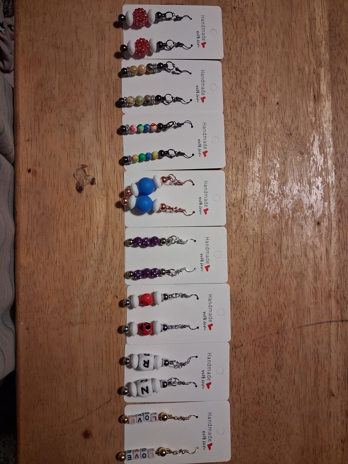 Pairs of colorful beaded drop earrings on white display cards, with patterned, letter, and sparkling beads