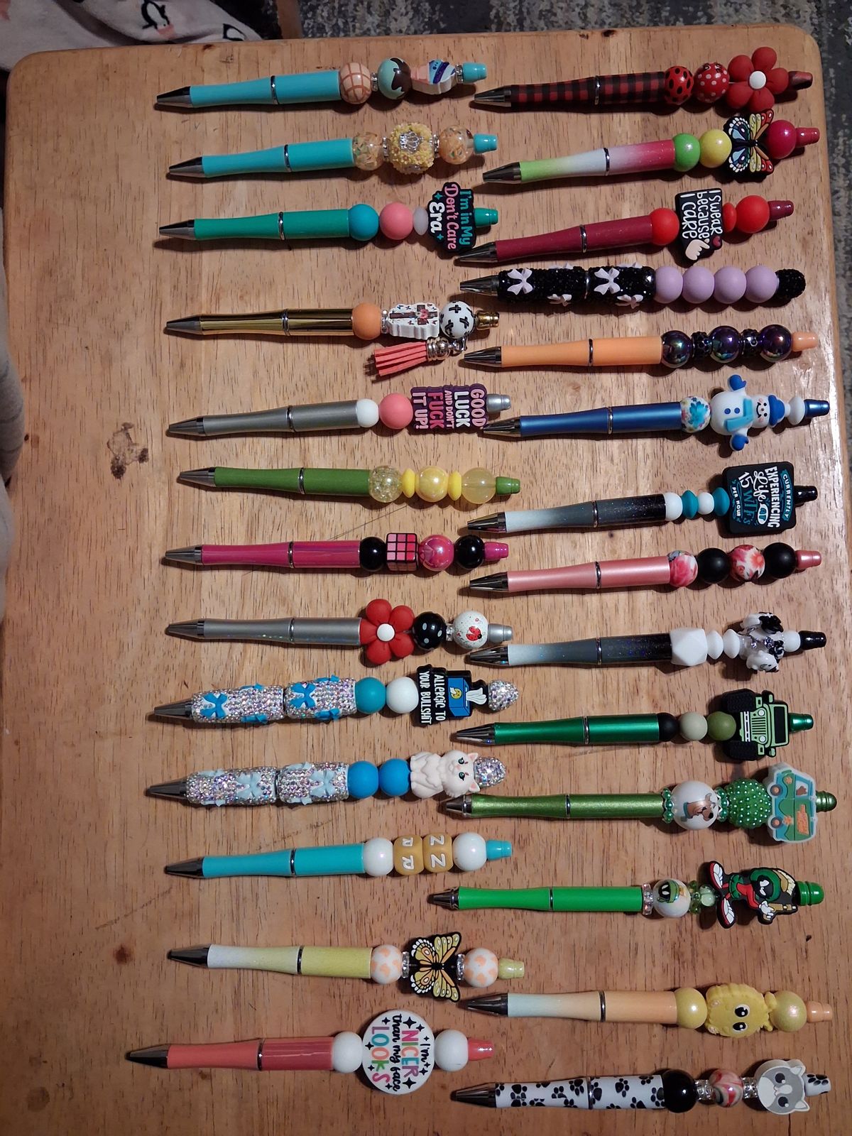 Colorful pens with decorative beaded tops, flowers, butterflies, animal charms, and sparkling details arranged on a wooden board