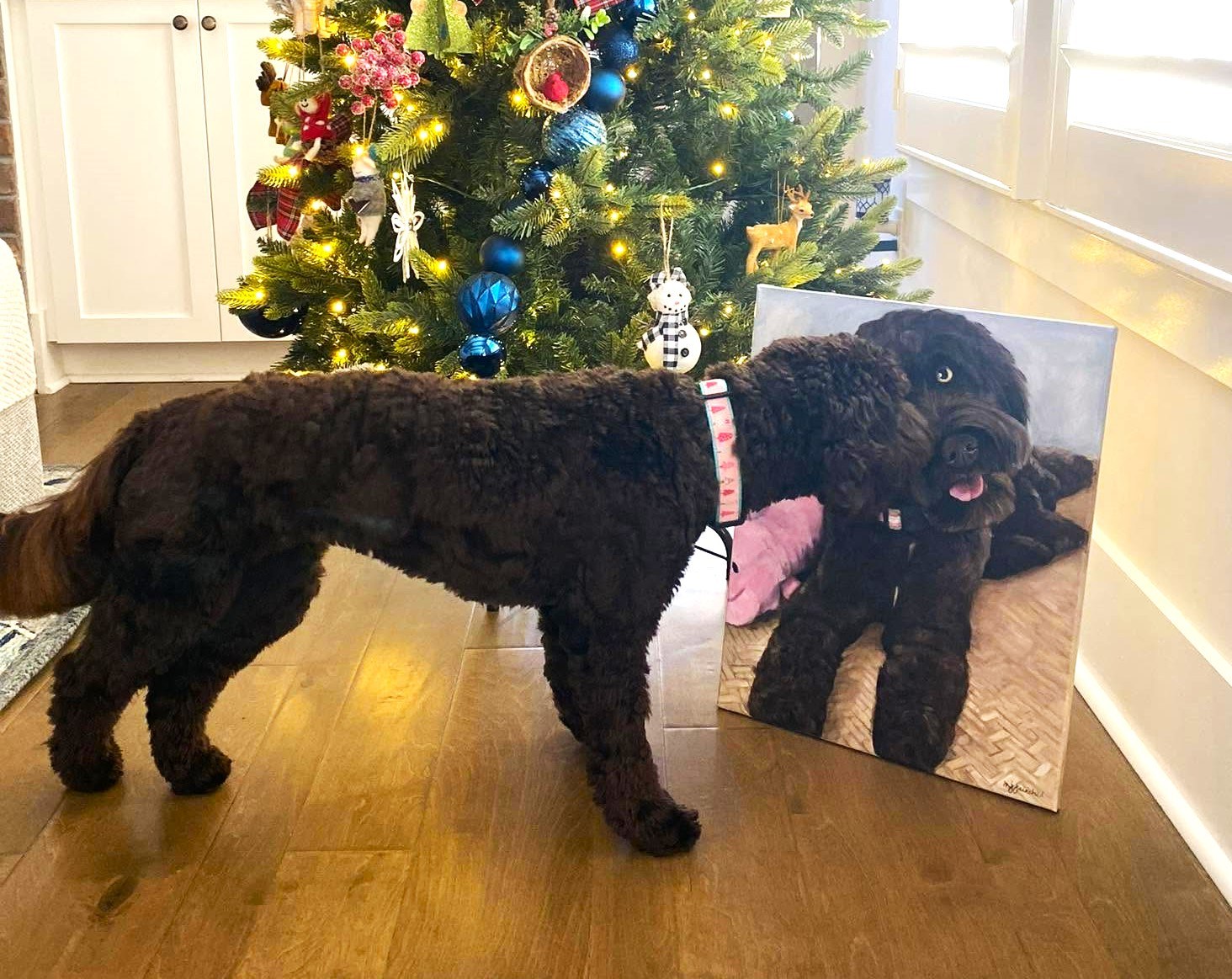 Black doodle sitting beside a painted pet portrait