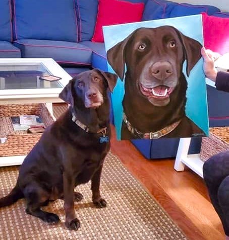 Chocolate Labrador sitting beside a painted pet portrait