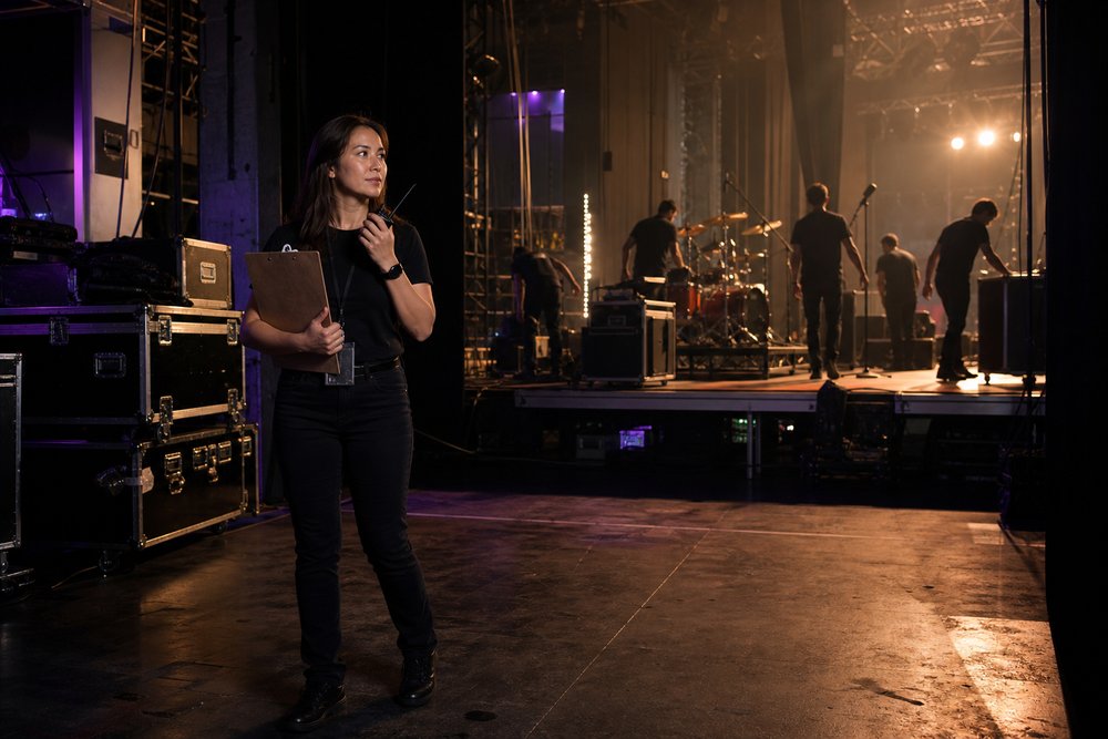 A tour coordinator holding a clipboard and radio backstage while stage crew prepare a concert venue