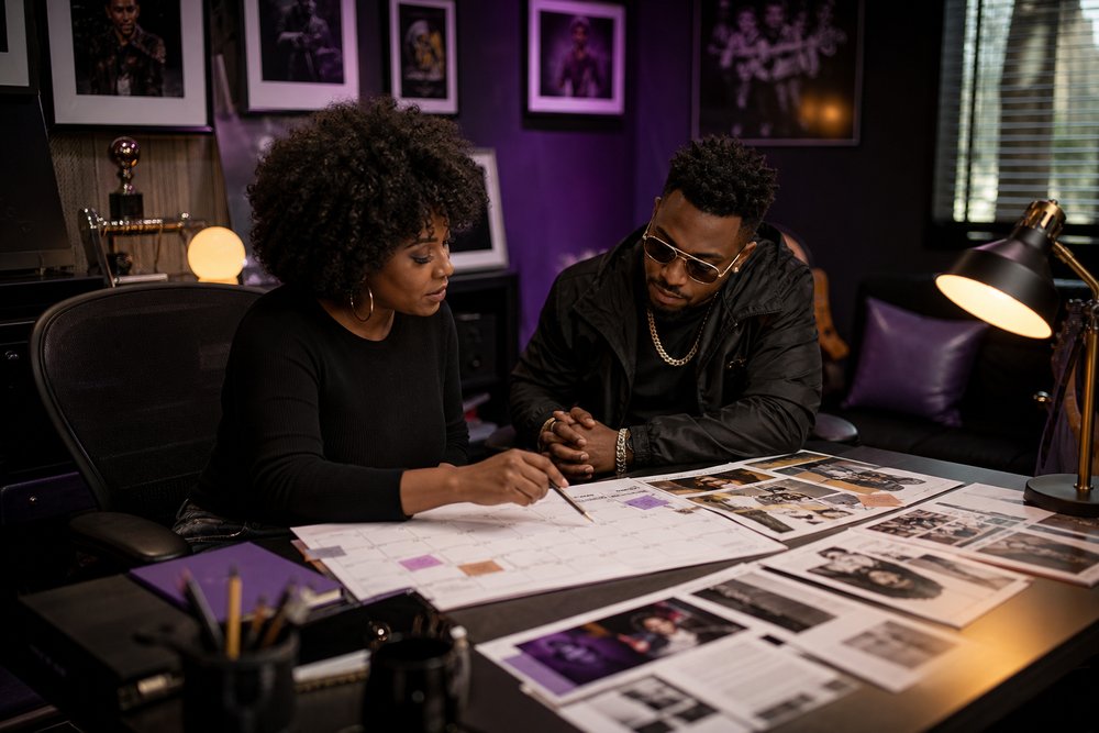 A Black publicist and recording artist discussing publicity plans with a calendar and press clippings in a creative office