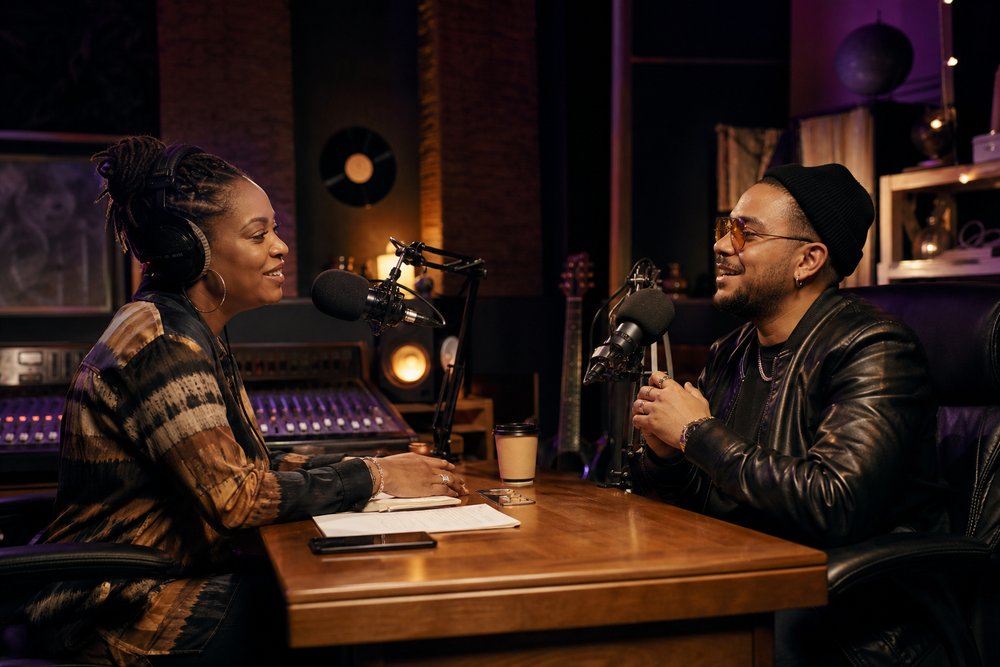 A Black female radio host interviewing a male musician across two broadcast microphones in a warmly lit recording room