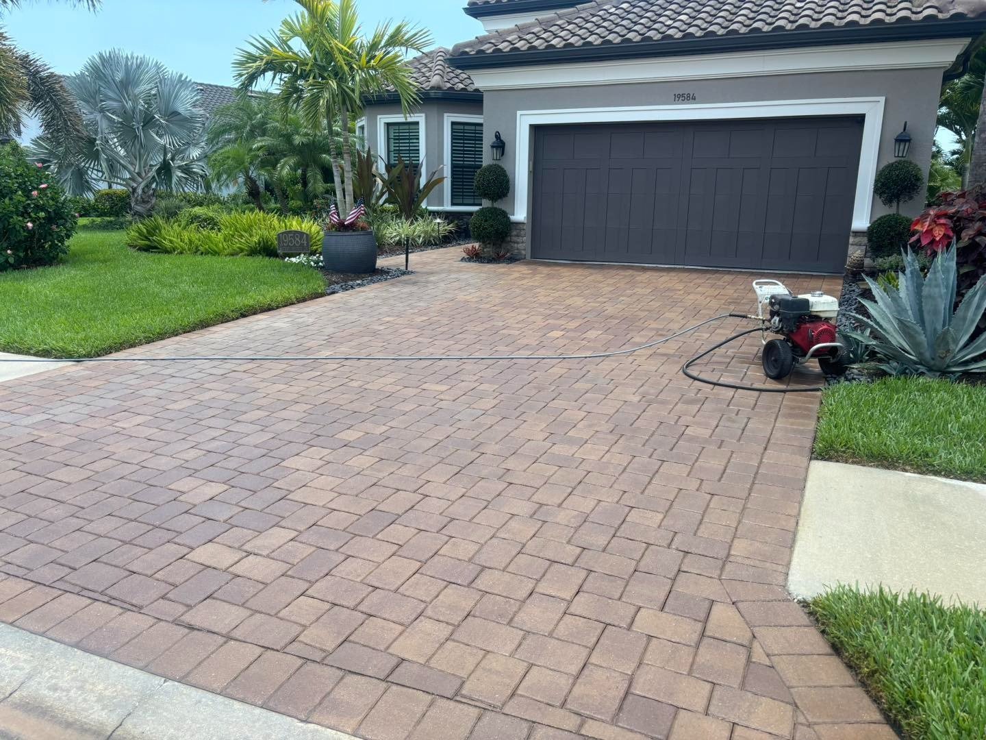 Brown paver driveway leading to a dark garage beside tropical landscaping