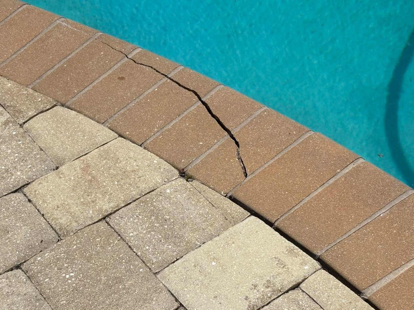 Cracked brown pool coping with an open joint beside pale deck pavers