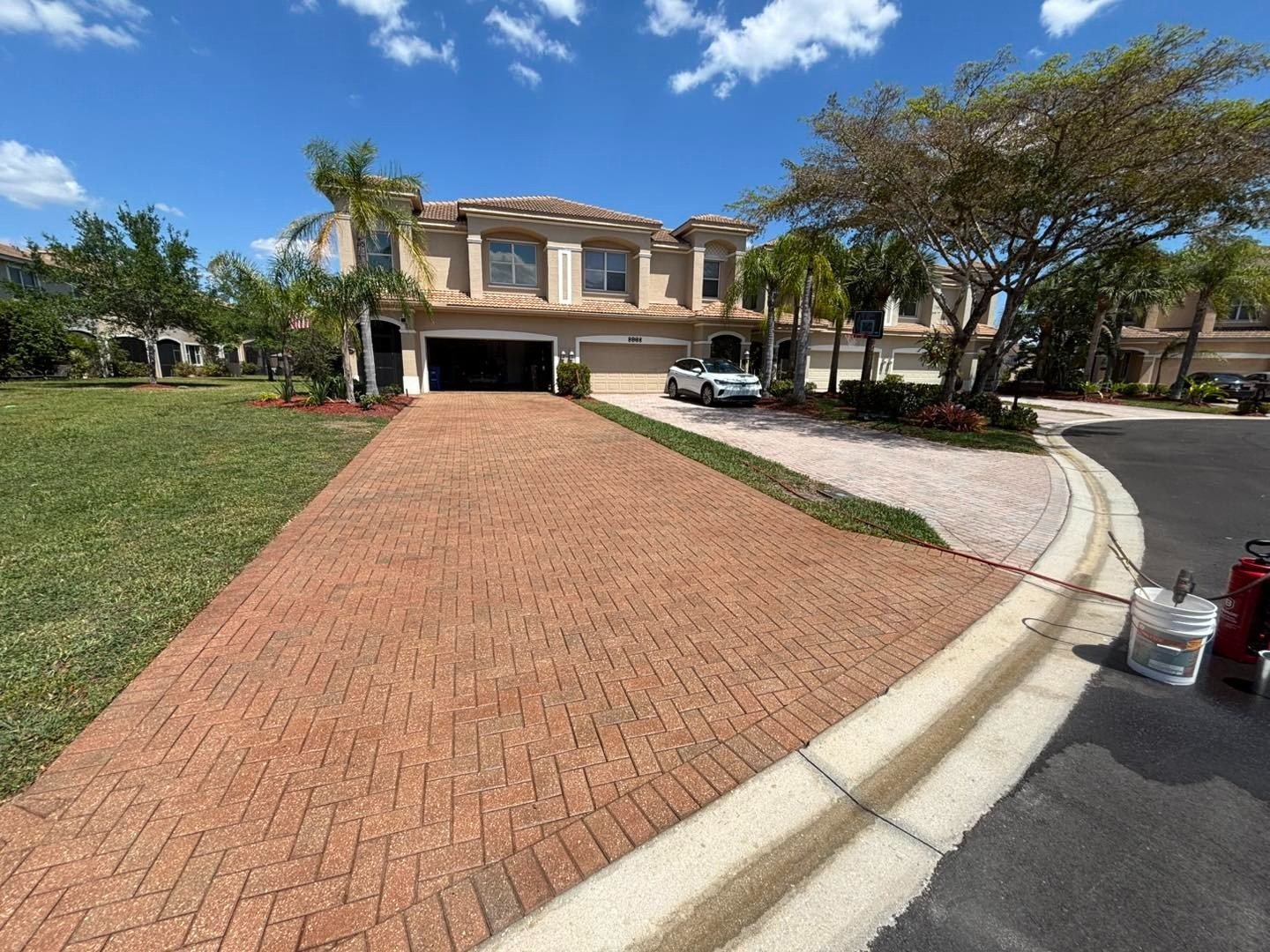 Long red herringbone paver driveway approaching a two-story home with palms