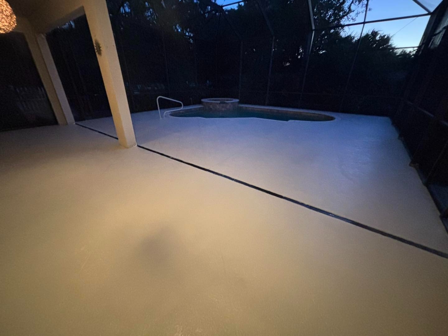 Pale pool deck at dusk beneath a screened enclosure beside a yellow house