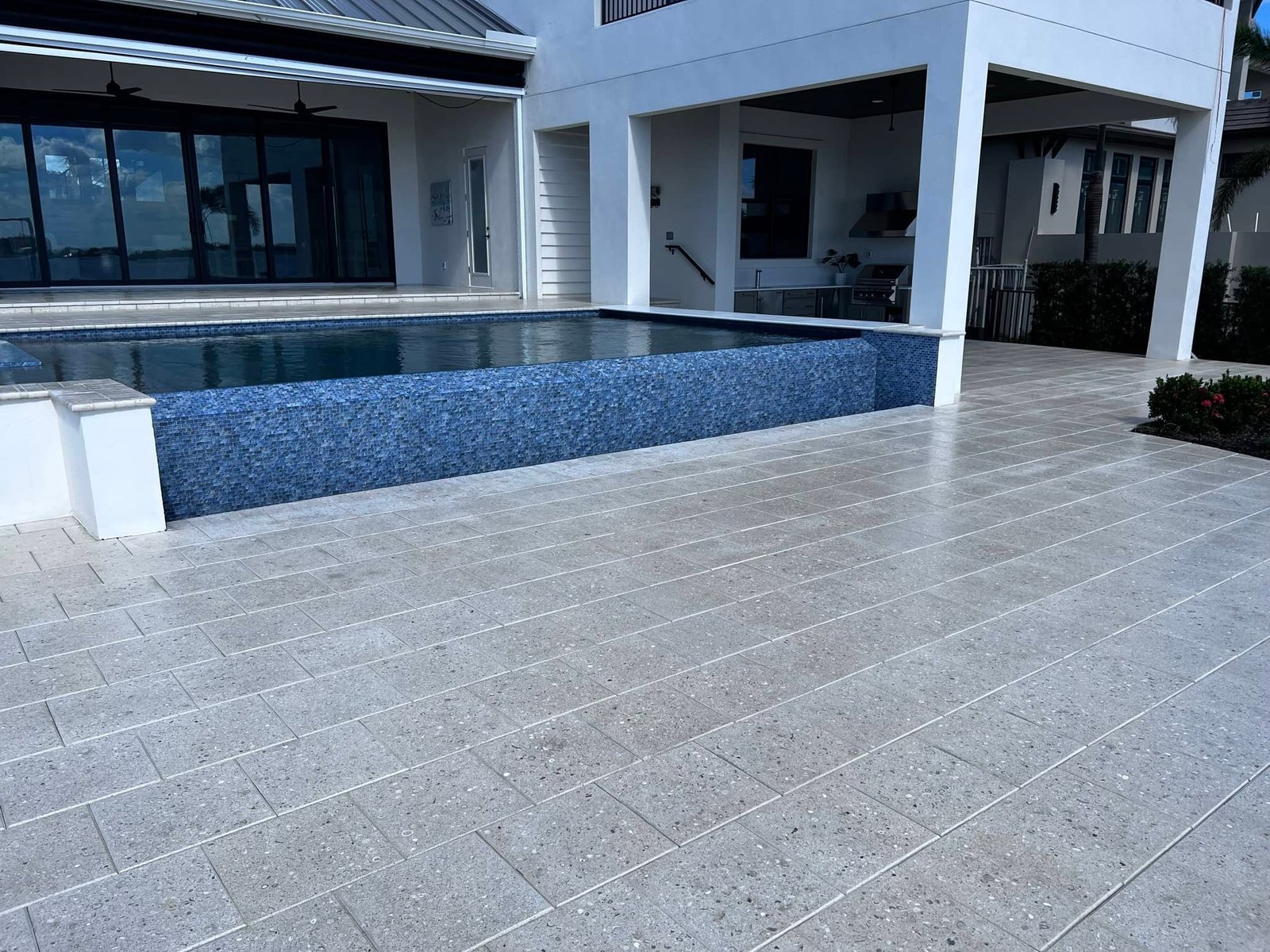 Pale rectangular poolside paving beside a blue-tiled pool and covered patio