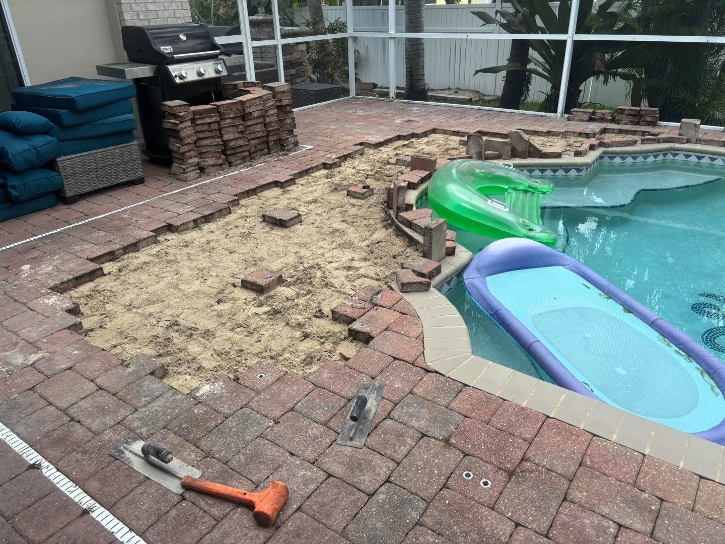 Pool deck opened to expose sand beside stacked pavers and pool floats