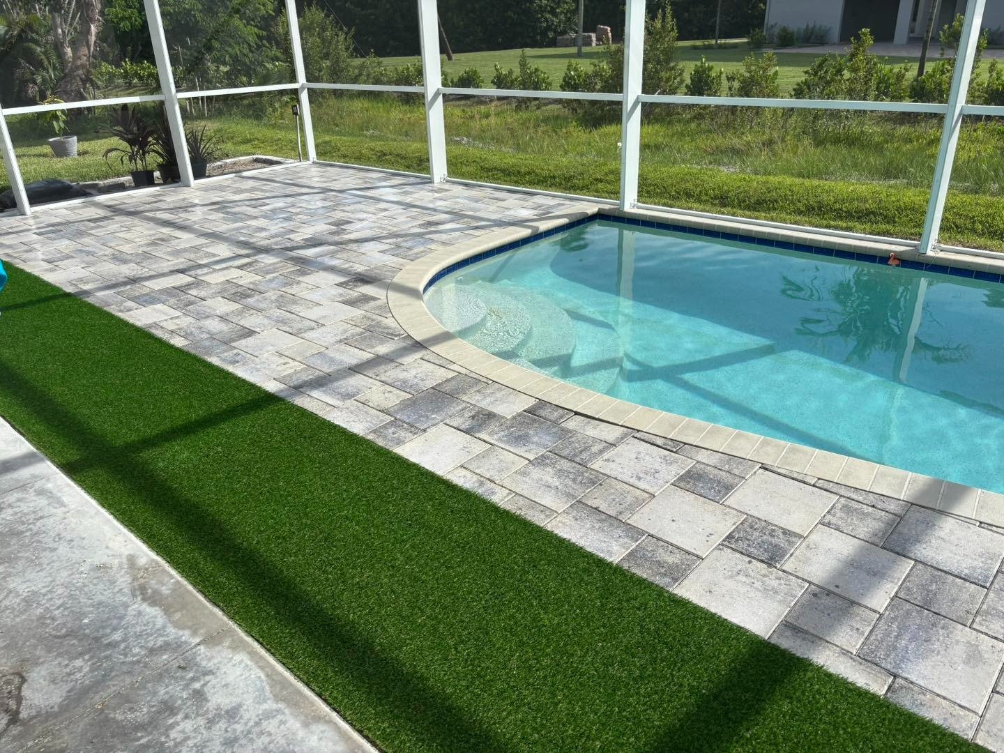 Screened swimming pool bordered by pale pavers and a bright green turf strip