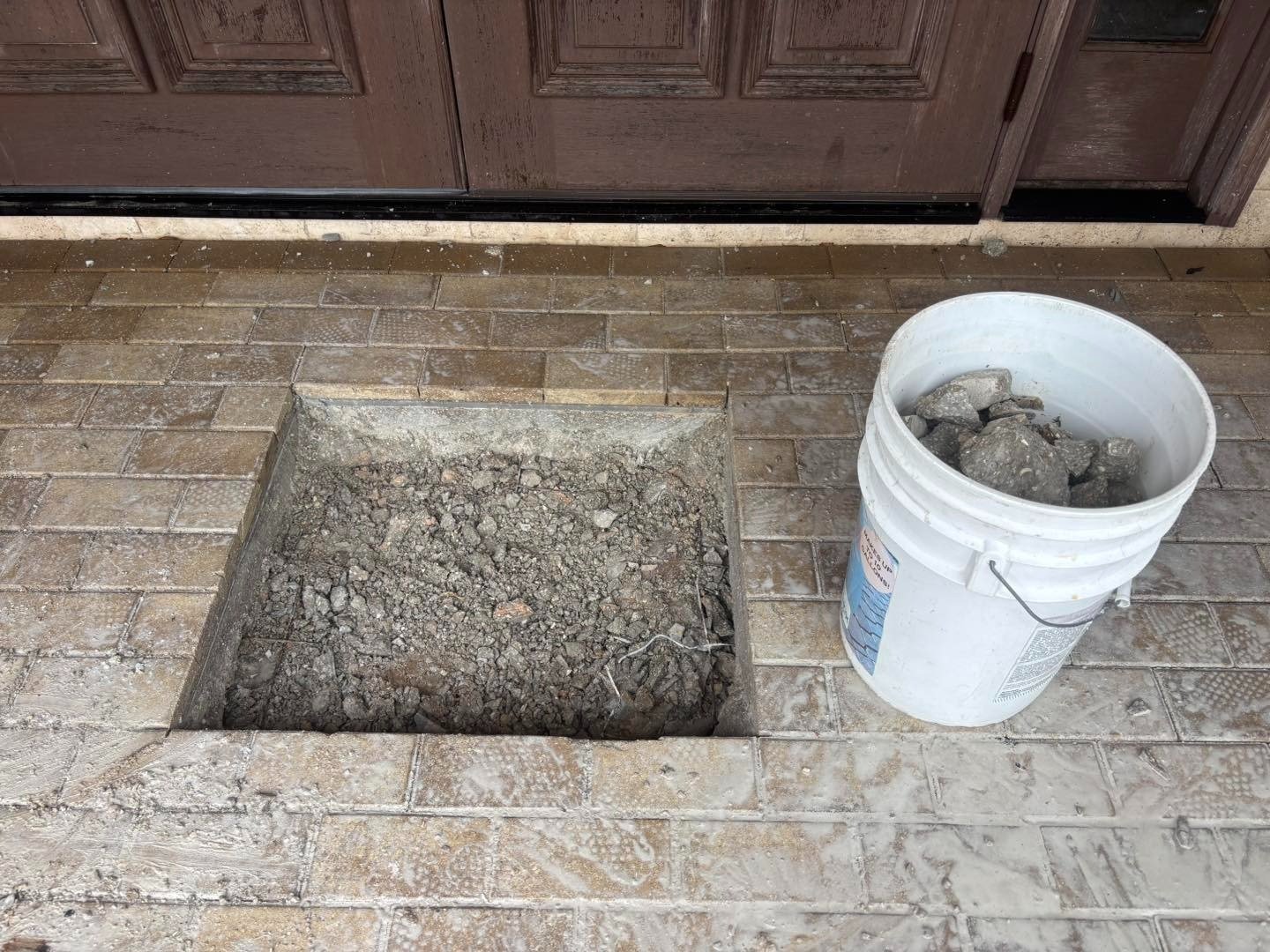 Square opening in an entryway paver surface beside a bucket of rubble