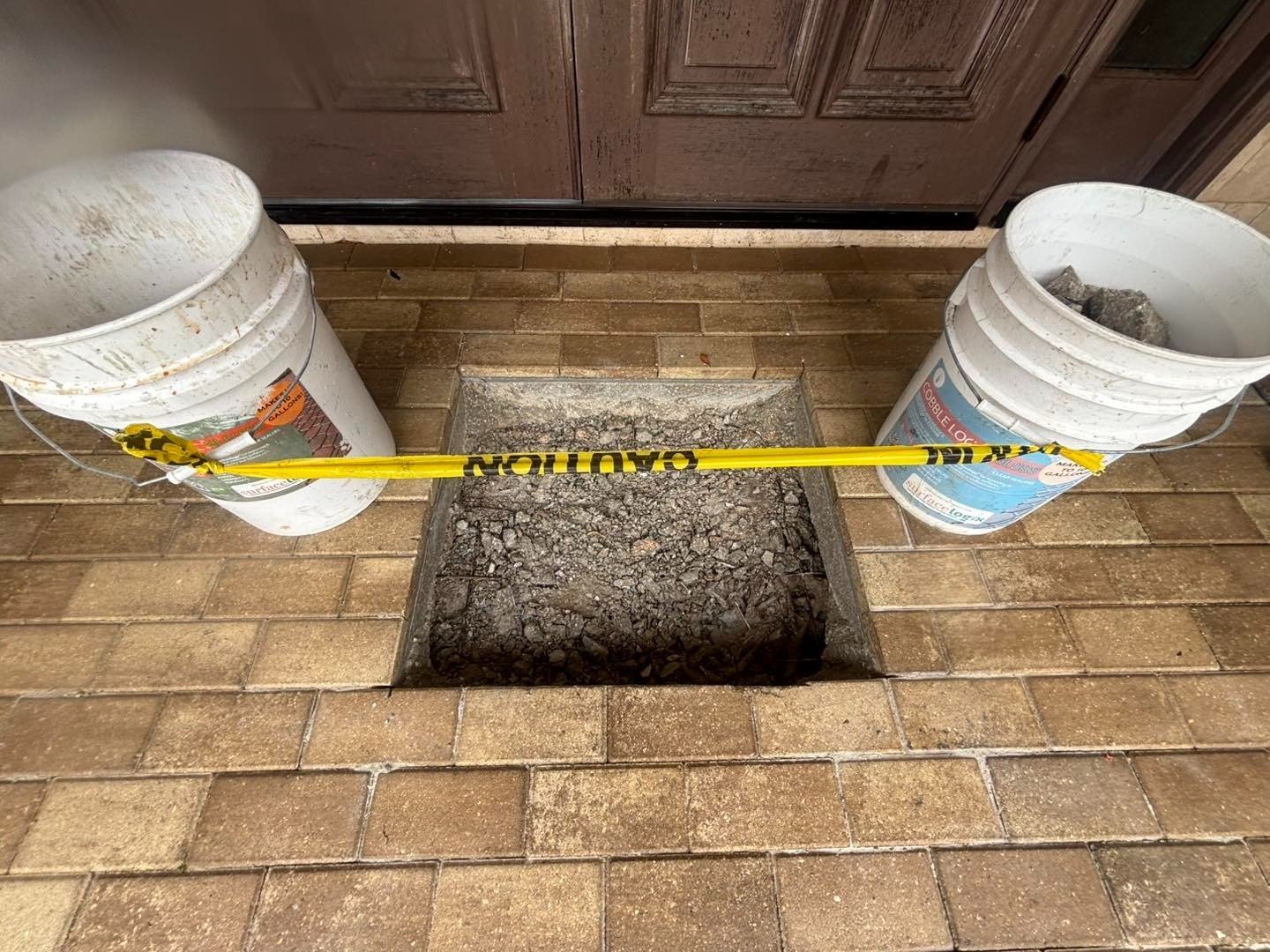 Square opening in paver entry blocked with caution tape between two buckets