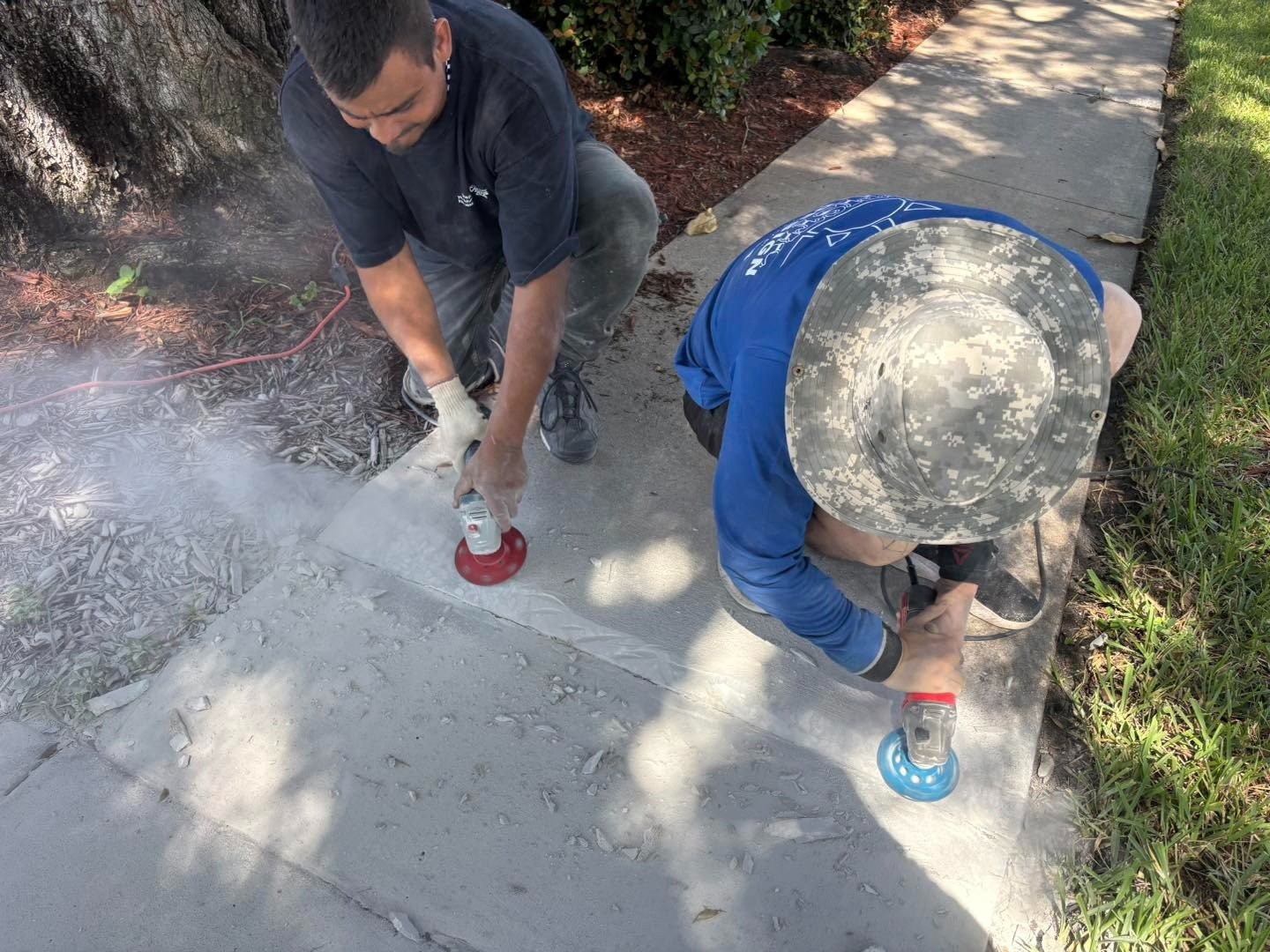 Two workers using handheld tools along a dusty concrete sidewalk joint