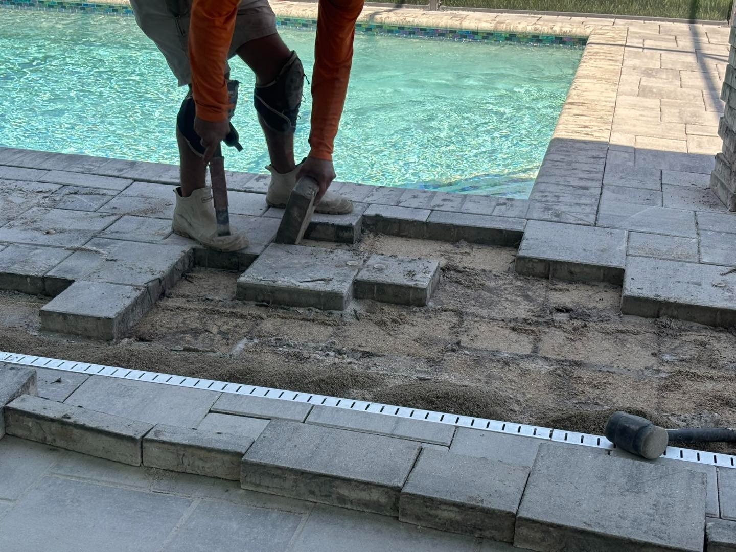 Worker lifting a paver beside an exposed pool deck base and measuring tape