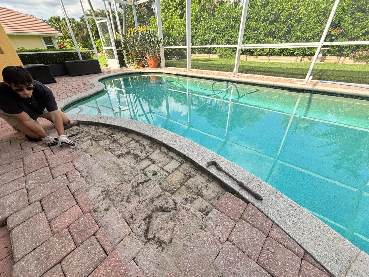 Worker lifting pavers beside a screened swimming pool with exposed ground underneath