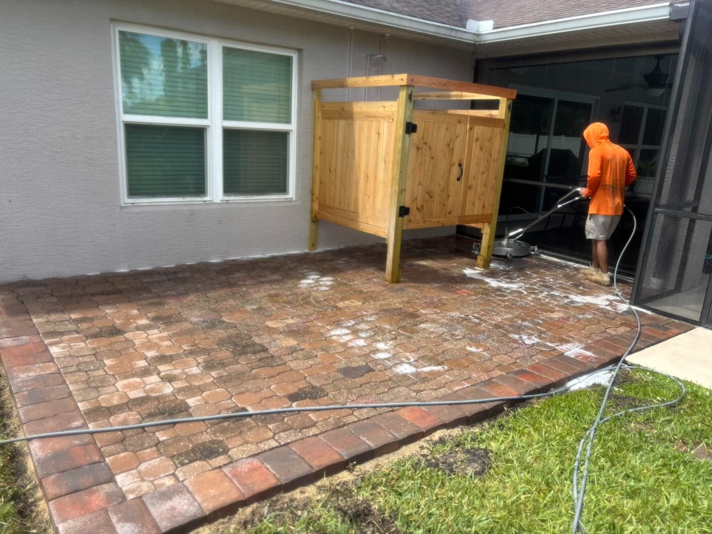 Worker washing a reddish paver patio beside a wooden outdoor shower enclosure