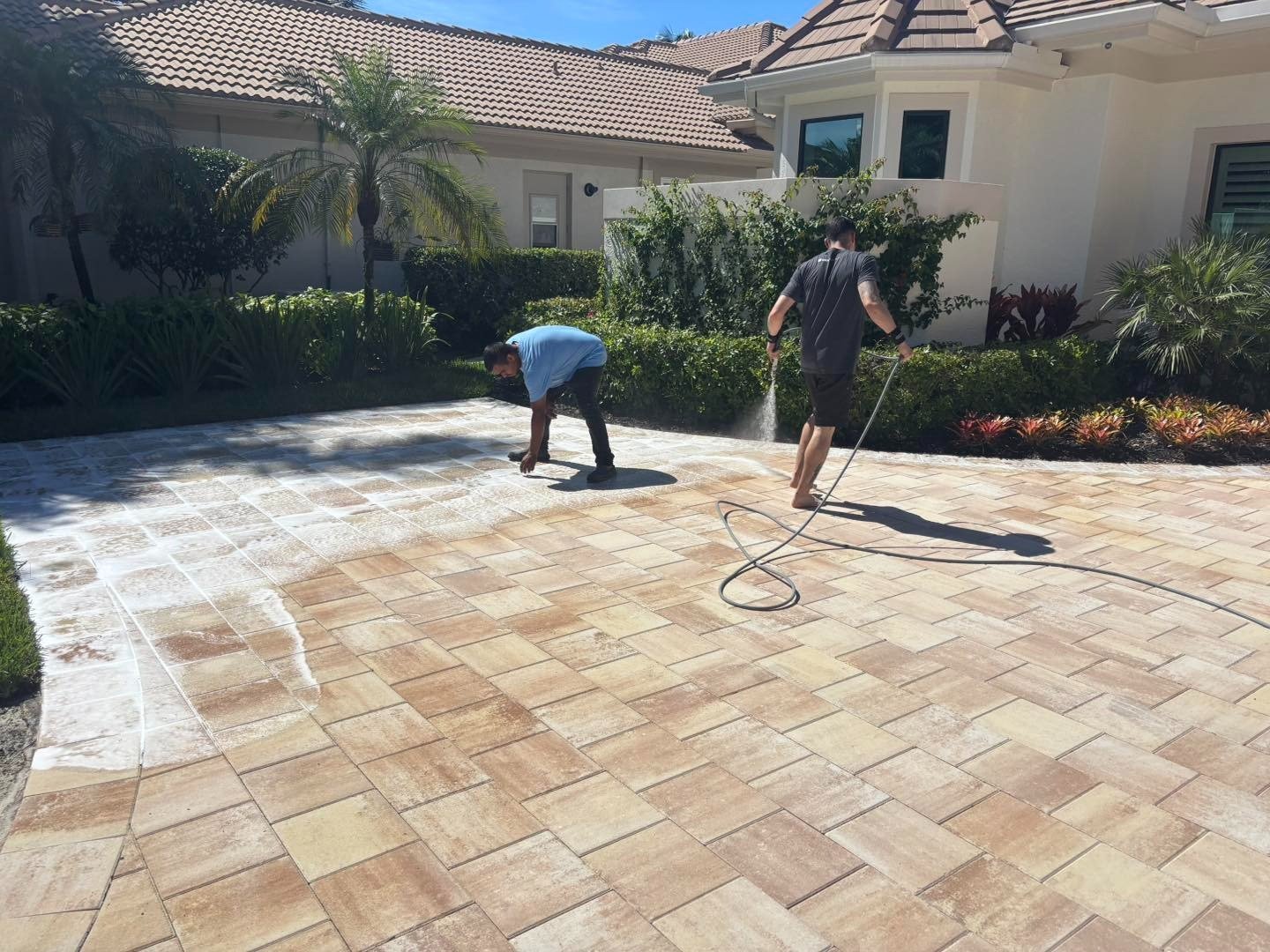 Two workers washing a broad multicolored paver driveway outside a house