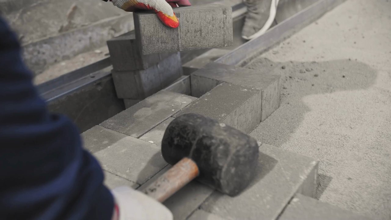 Gloved hands positioning a gray paving block beside a rubber mallet