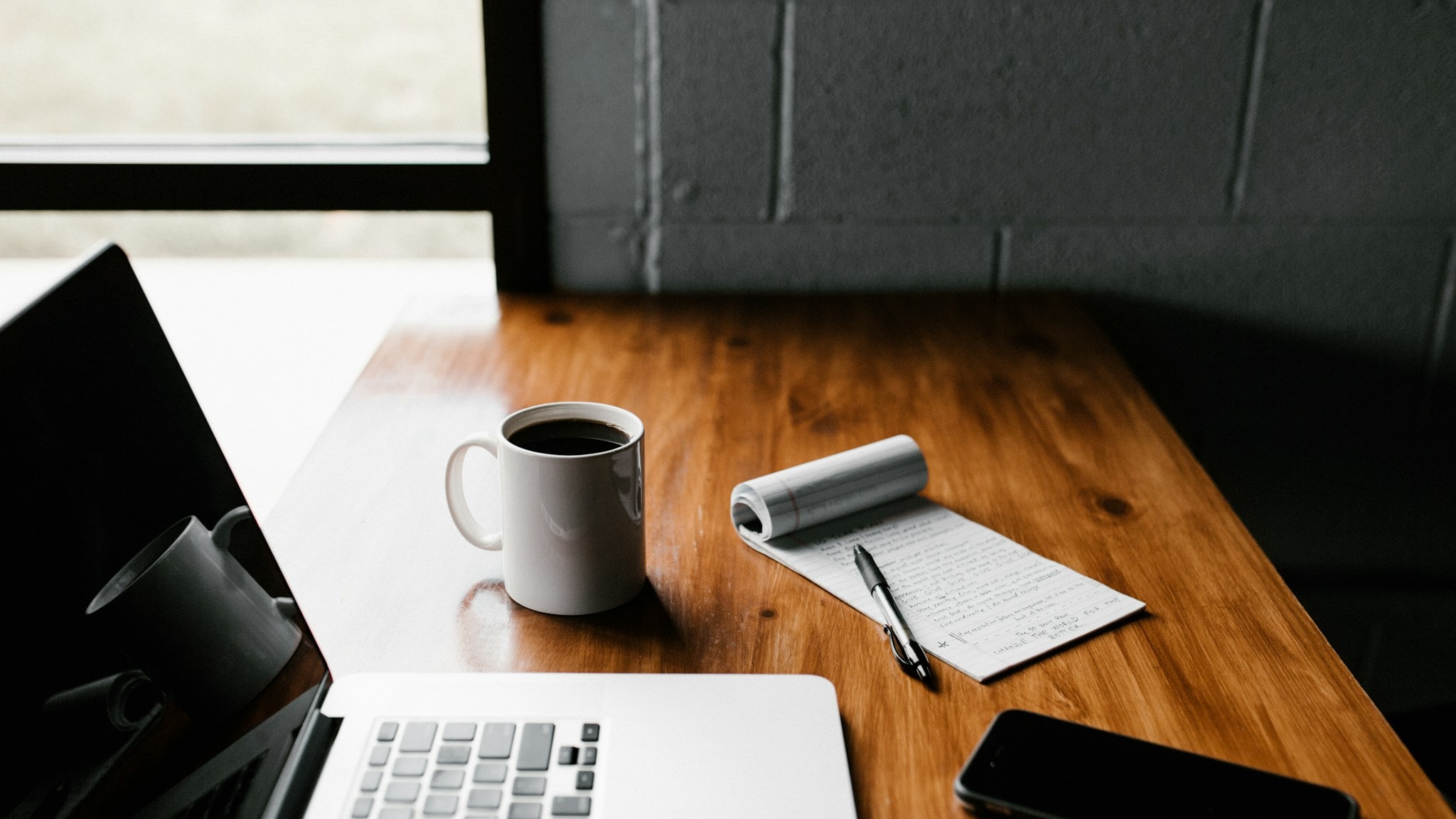 Laptop, notebook, coffee, and phone arranged on a wooden desk
