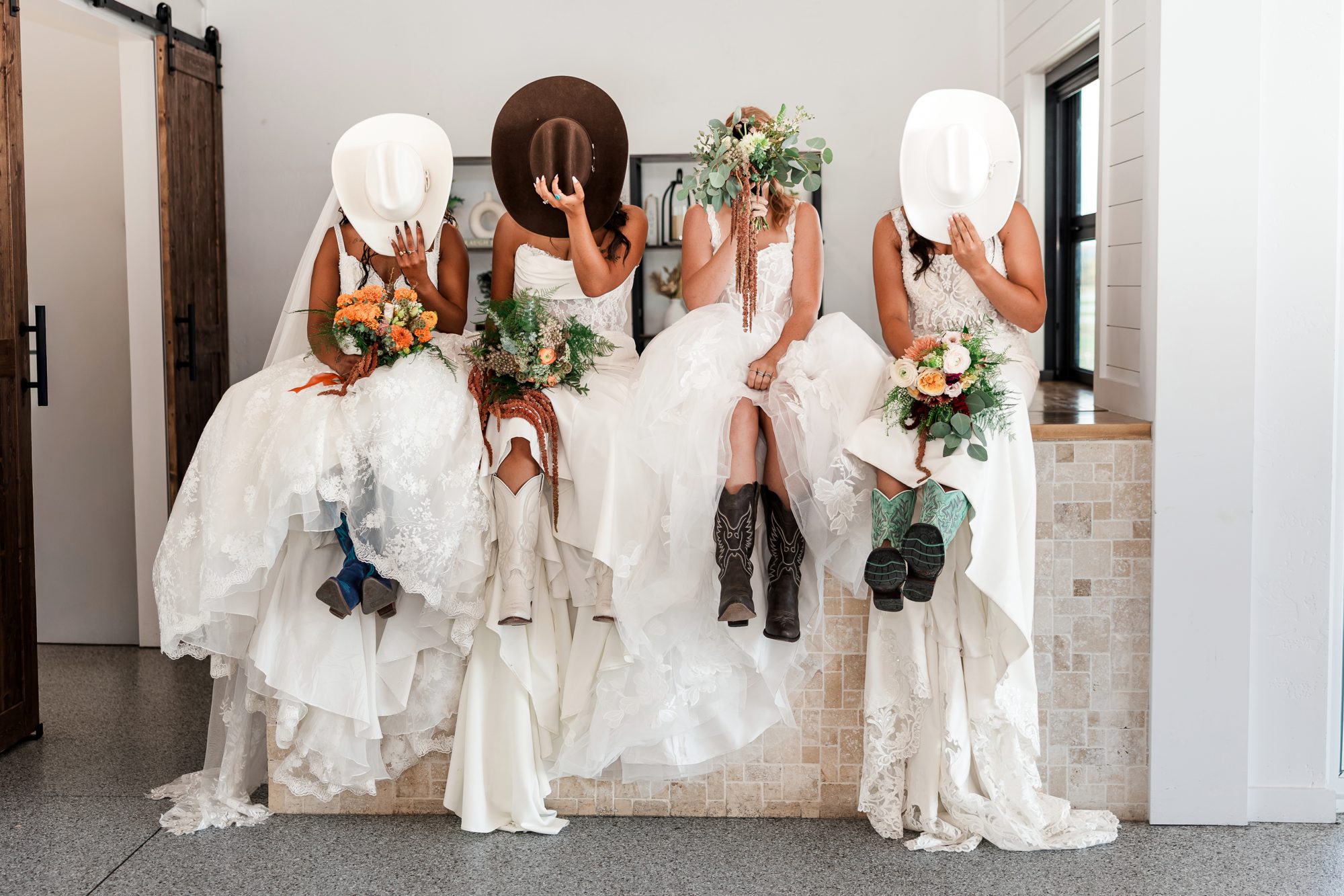 Four brides sitting together in wedding gowns, holding hats and bouquets in front of their faces