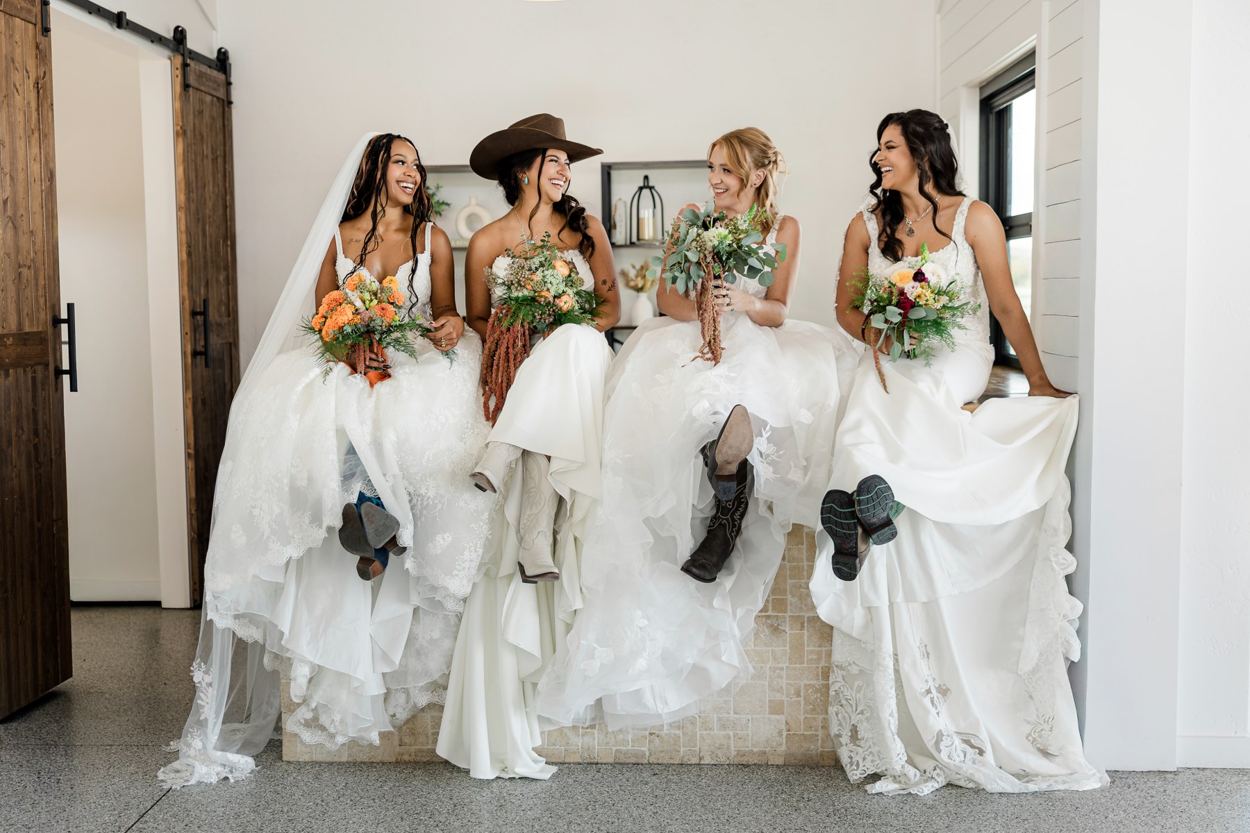Four brides laughing together in different wedding gown styles, holding bouquets and showing their boots