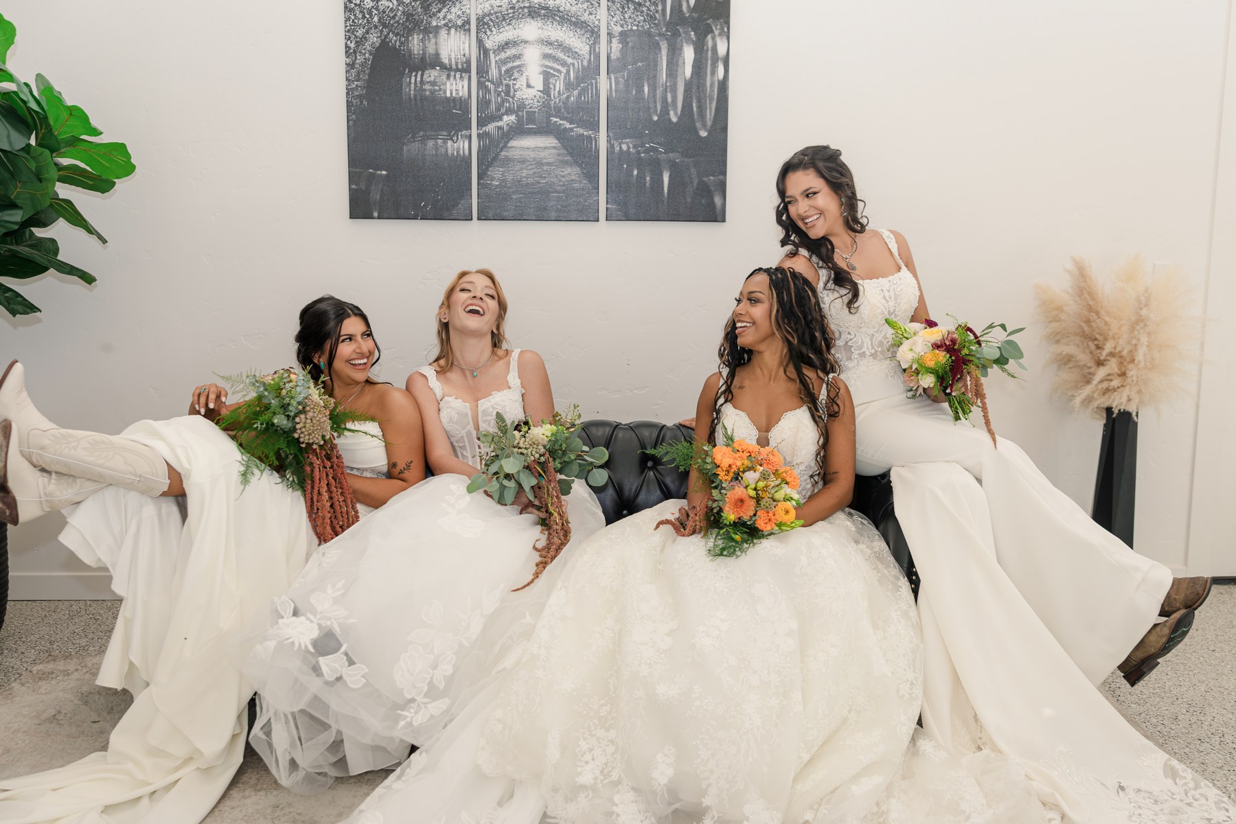 Four brides laughing together on a sofa in their wedding gowns, holding colourful bouquets