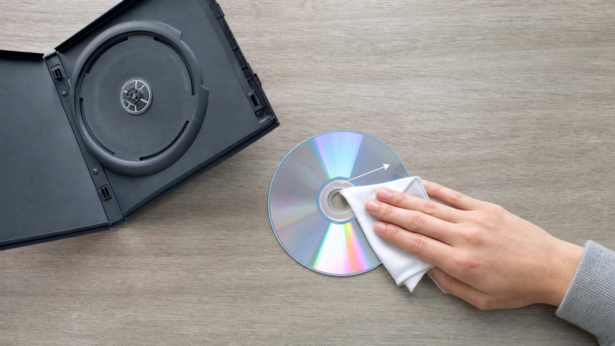 A hand wiping an unbranded DVD in a straight line from the center toward the outer edge with a soft cloth