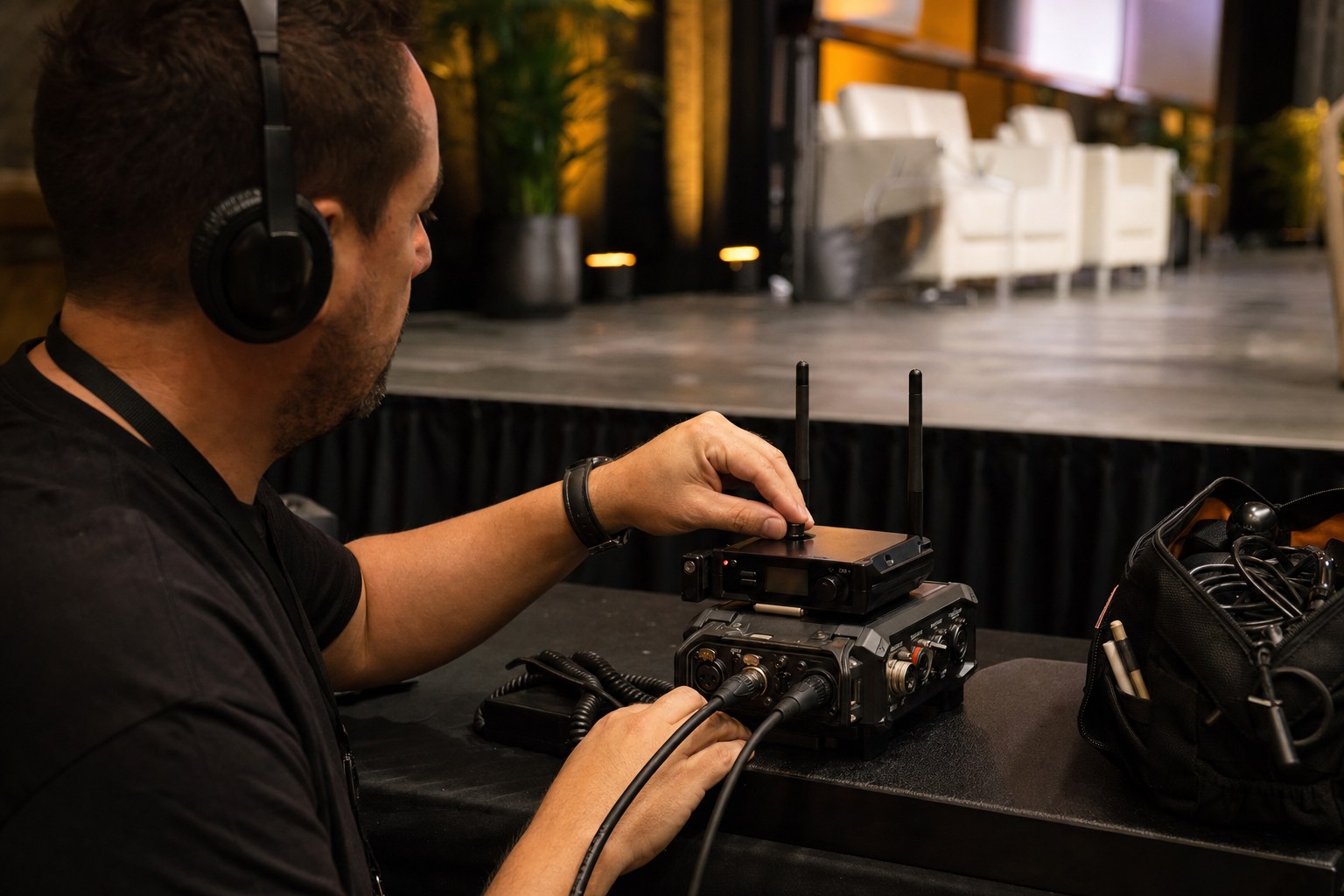 Audio technician checking a field recorder and wireless microphones beside a conference stage