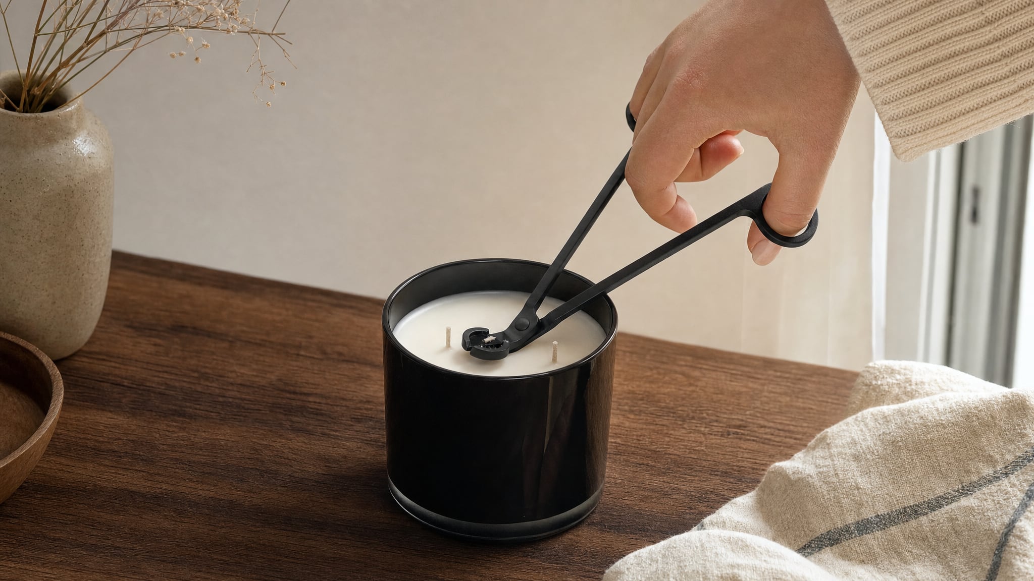 Hands using a wick trimmer on a cooled black container candle.