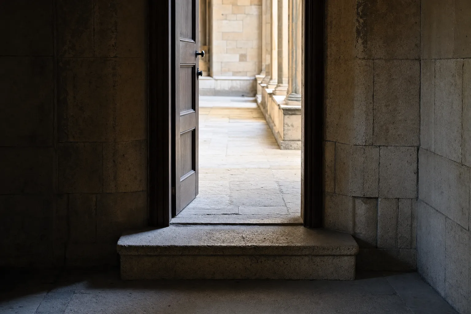 A single stone step leading through an open wooden doorway toward a sunlit stone corridor
