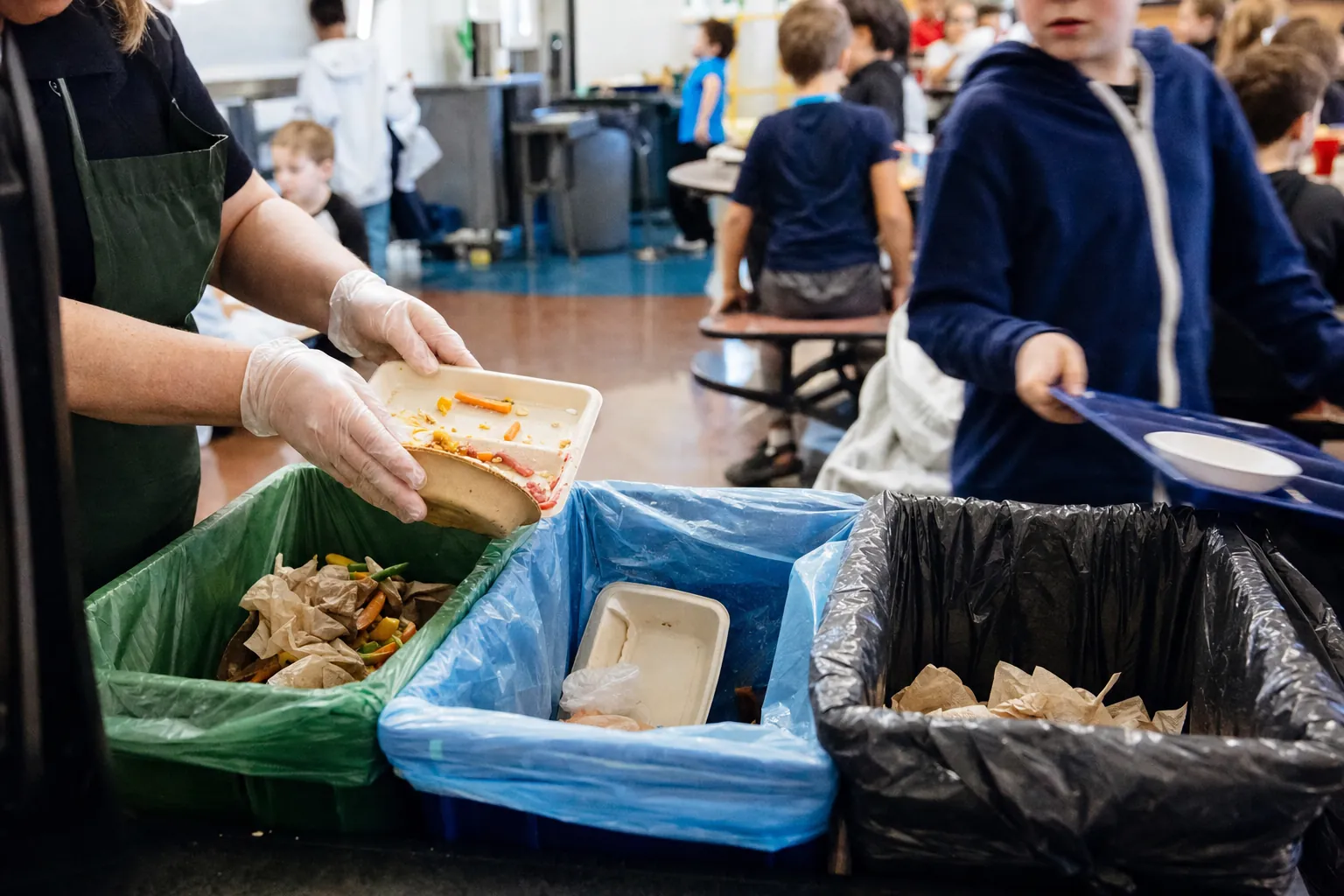 Lunchroom staff sorting meal packaging beside waste bins.