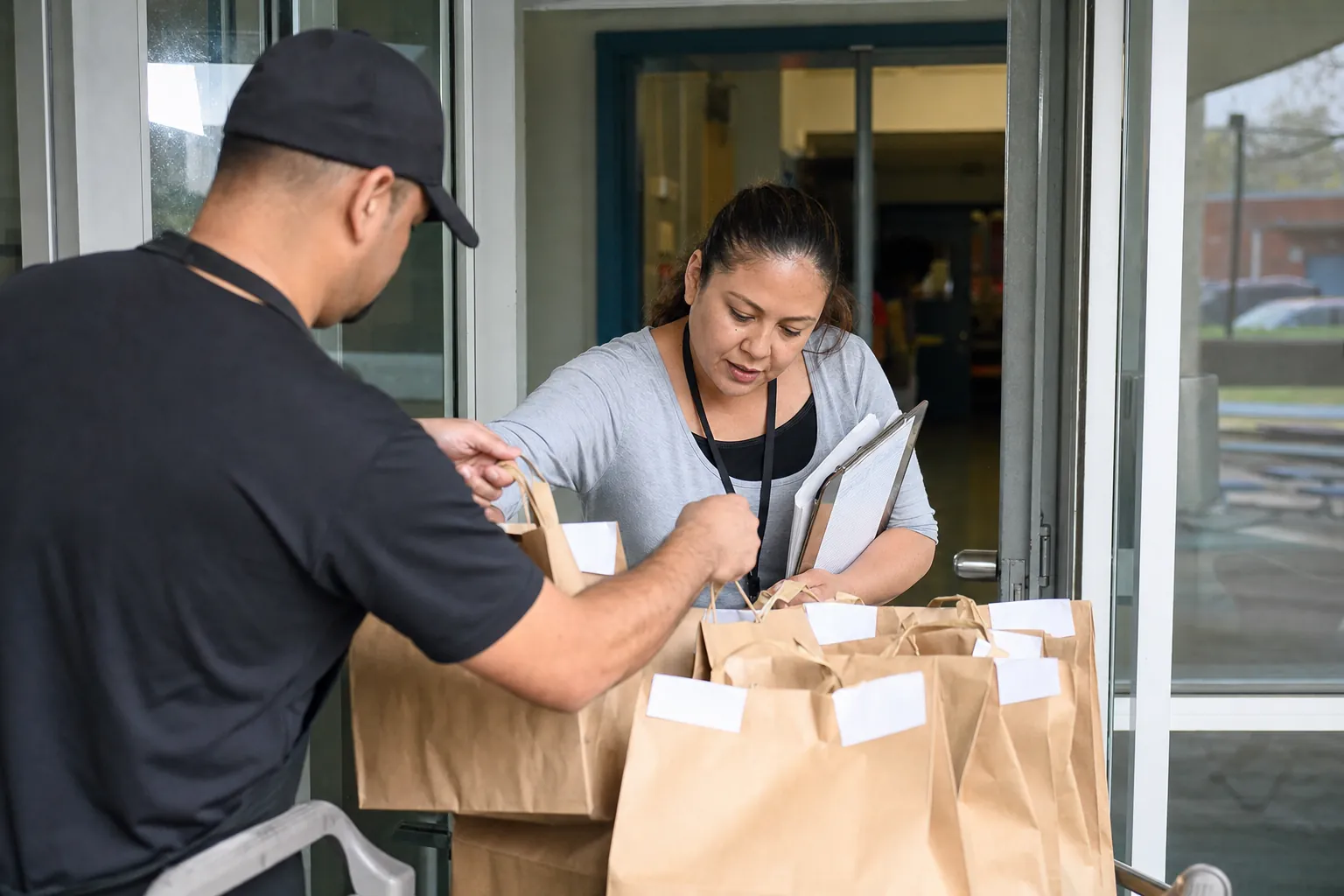 School staff member receiving labeled restaurant lunch bags from a delivery partner at a school entrance.