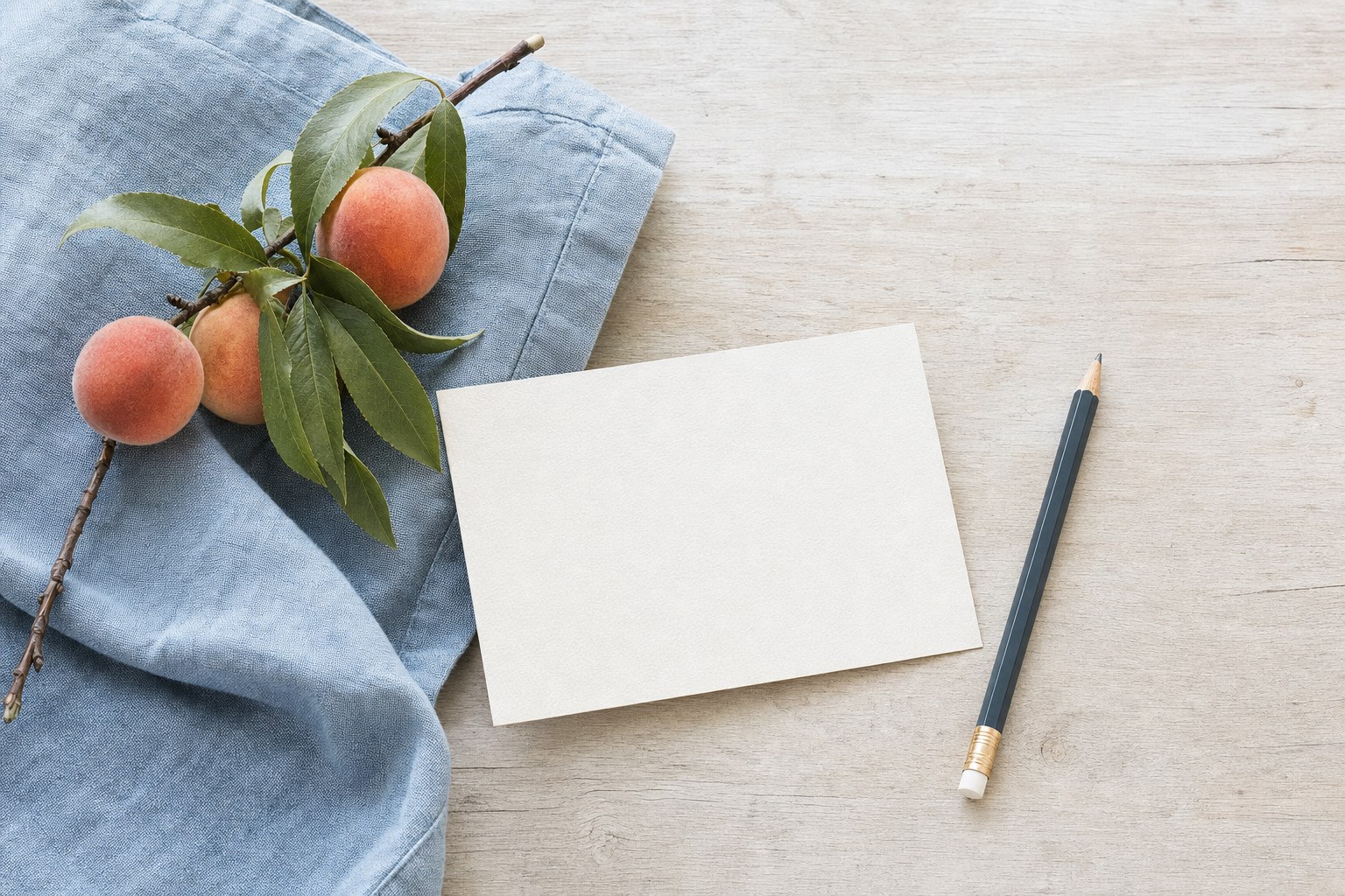 A blank note card, pencil, peach branch, and blue linen on a pale wood table