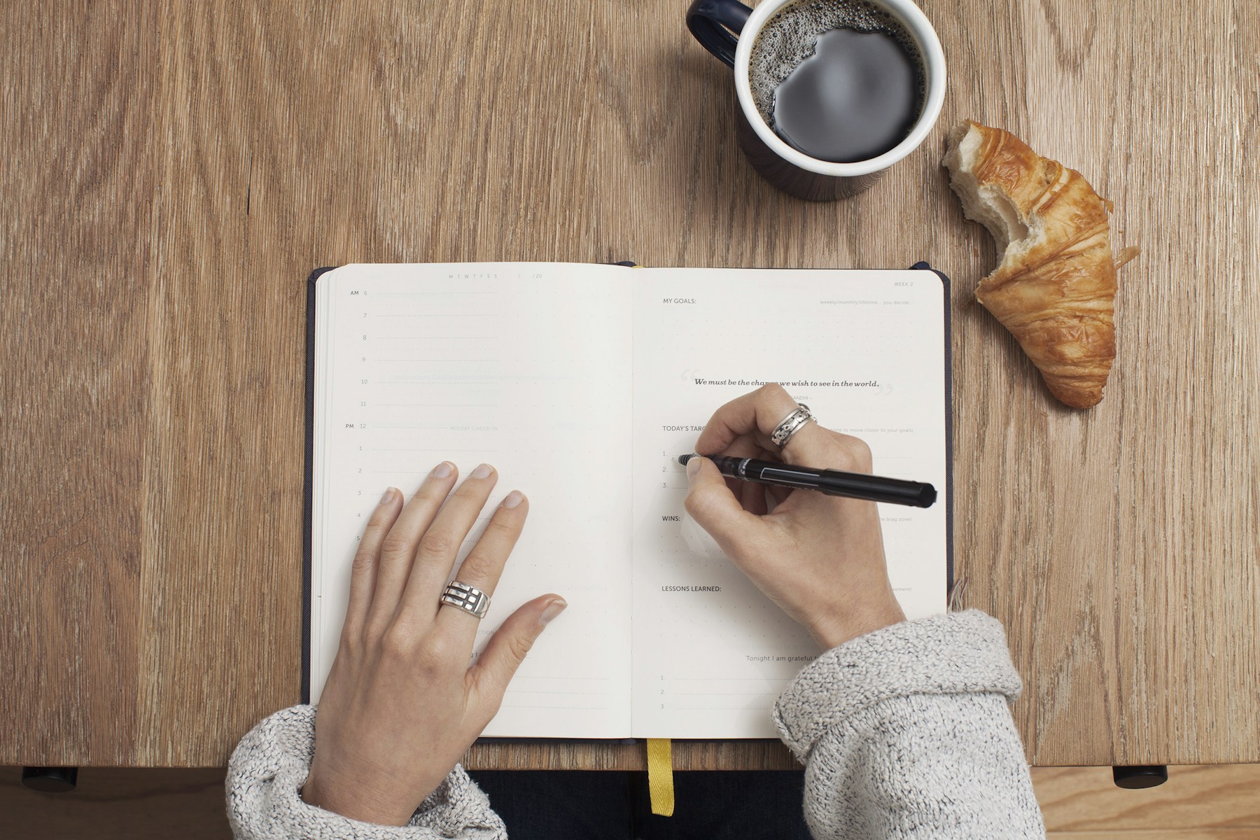 A person writing in an open planner beside coffee and a pastry on a wooden table