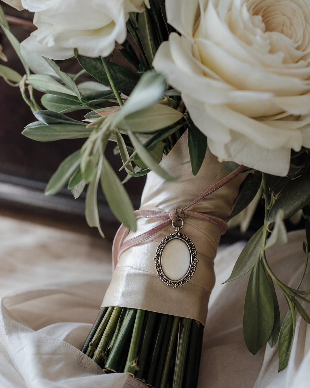 Blank oval memory charm tied to an ivory bridal bouquet with white roses and olive leaves