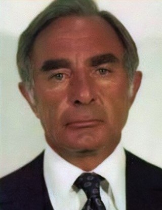 A close-up photo of an older man with gray hair, wearing a dark suit and white shirt with a patterned tie, looking directly at the camera.