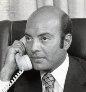 A man speaking on a vintage telephone, wearing a suit and tie, with a serious expression.