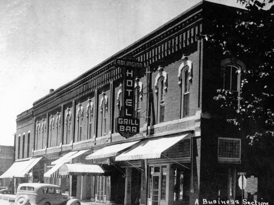 Historic black-and-white photo of the Arlington Hotel featuring a prominent sign that reads 'Arlington Hotel Grill Bar,' with a view of the brick building and nearby vintage cars.