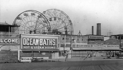 Historic black and white photograph of a seaside amusement area featuring the Ocean Baths building, a large sign advertising hot and steam rooms, and a ferris wheel in the background.