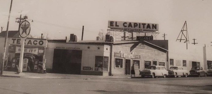 Black and white photograph of a street scene featuring a Texaco gas station on the left and a restaurant named El Capitan on the right, with classic cars parked in front.