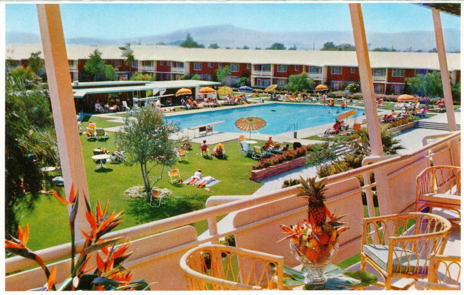 A vibrant resort pool area with people lounging by the pool, sun umbrellas, and lush greenery. In the foreground, there are colorful chairs and a decorative fruit bowl.