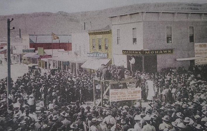 Historical black and white image of a large crowd gathered in a town square, with a speaker on a platform addressing the audience, several buildings and storefronts visible in the background.