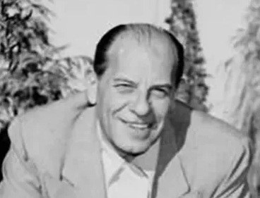 A smiling man in a light-colored suit, posing outdoors with greenery in the background.
