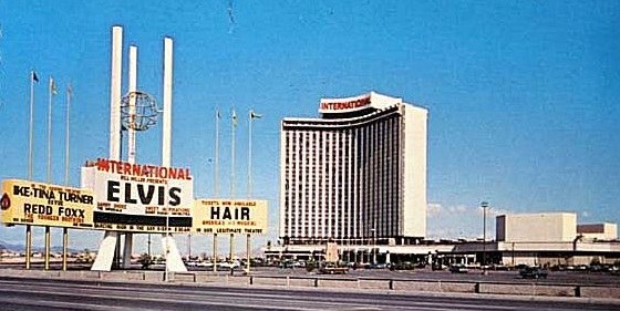A vintage photograph of the International Hotel in Las Vegas, featuring a prominent sign advertising performances by Elvis Presley, Tina Turner, and Redd Foxx, with clear blue skies in the background.