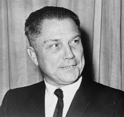 Black and white portrait of a man in a suit, looking to the side with a slight smile, against a backdrop of draped fabric.